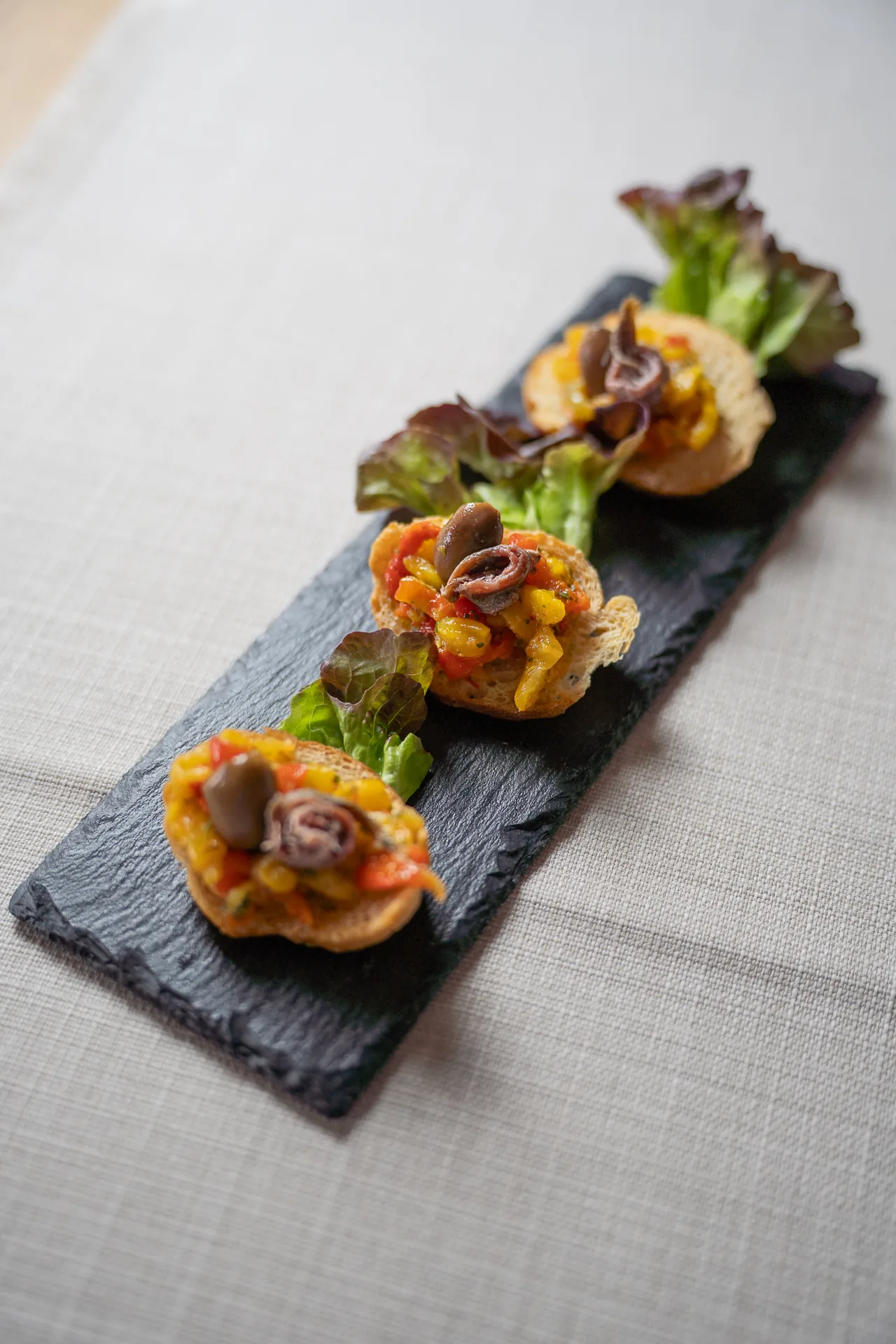 Tre crostini con verdure, olive e alici su un vassoio di ardesia.